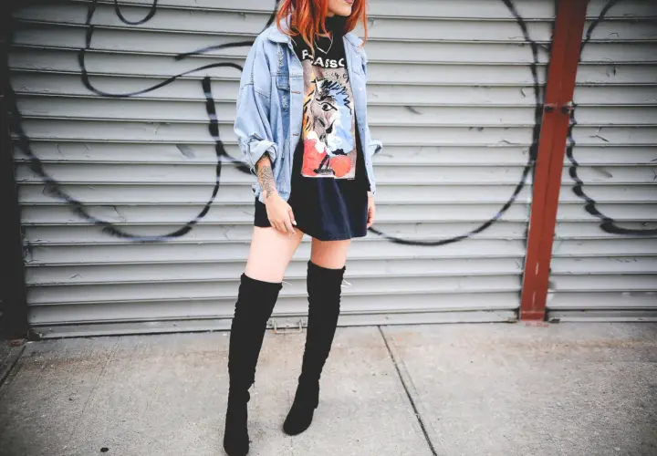 Chica con botas altas para bajitas, chaqueta vaquera y camiseta gráfica, posando frente a una pared de garaje con grafitis.
