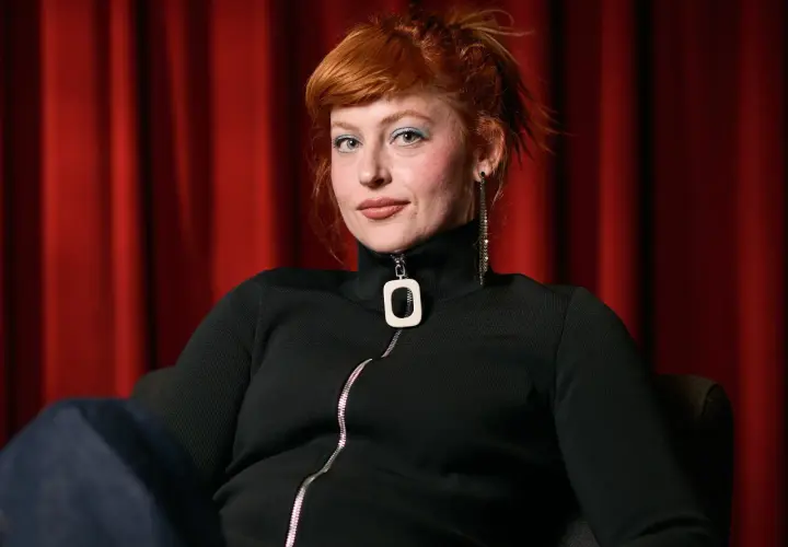 Chloe Wallace, con su cabello pelirrojo recogido y maquillaje azul, posa frente a una cortina roja.