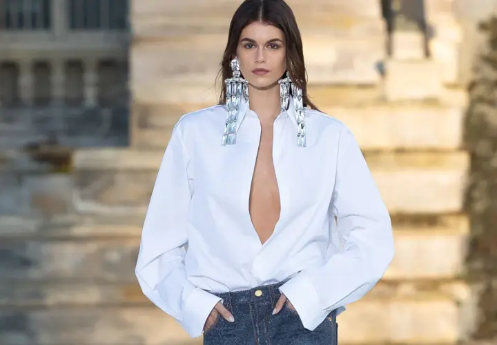 Mujer con camisa blanca desabrochada y vaqueros, luciendo grandes pendientes brillantes.