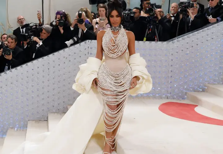 Kim Kardashian desciende la escalera de la Met Gala, deslumbrando con un vestido de perlas y una capa voluminosa, rodeada de fotógrafos.