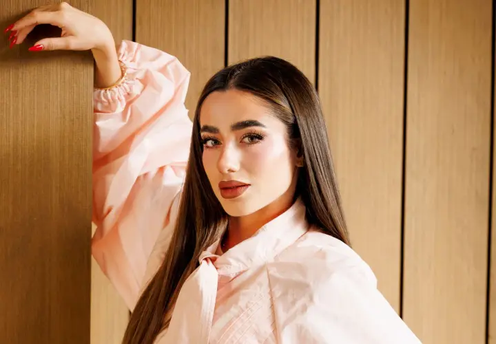 Lola Lolita, con su cabello largo y liso, posa con un vestido rosa pálido, apoyando su mano en un pilar de madera.