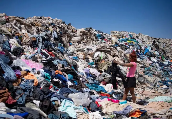 Montaña de ropa desechada, reflejo de las **fast fashion consecuencias**. Una joven busca entre los residuos textiles bajo un cielo azul.