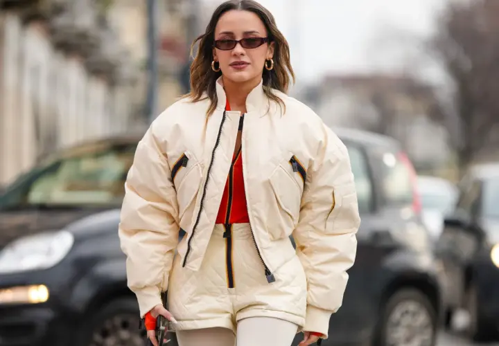 Mujer con gafas de sol y pendientes dorados, luciendo una bomber acolchada color crema sobre un top naranja y shorts a juego.