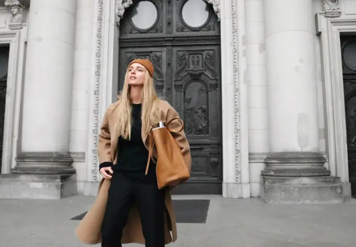Mujer con abrigo camel, gorro y bolso marrón, luciendo un elegante pantalon negro.