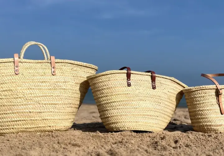 Tres capazos de playa grandes de palma, con asas de cuero, descansan en la arena bajo un cielo azul.