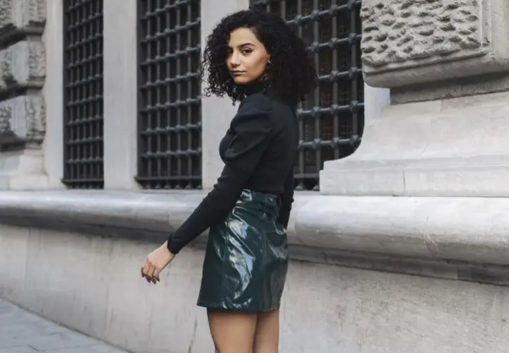 Joven con pelo rizado y falda verde brillante, estilo inspirado en Mary Quant, posa frente a un edificio histórico.