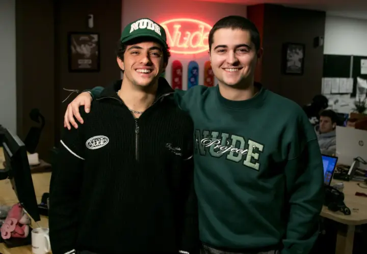Bruno Casanovas y su amigo sonríen, con un letrero de neón "Nude" detrás.