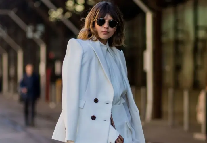 Mujer con traje blanco, gafas de sol redondas y cabello castaño. Su atuendo, elegante y sencillo, refleja el **minimalista significado** de la moda.