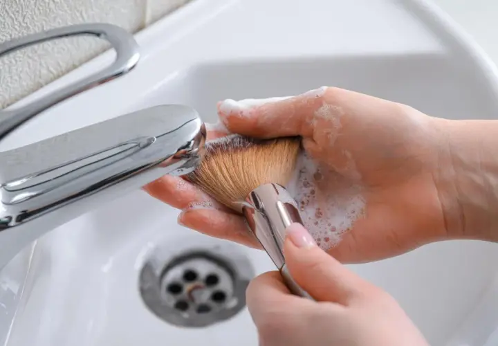Manos limpiando brochas de maquillaje con espuma y agua bajo un grifo cromado en un lavabo blanco.