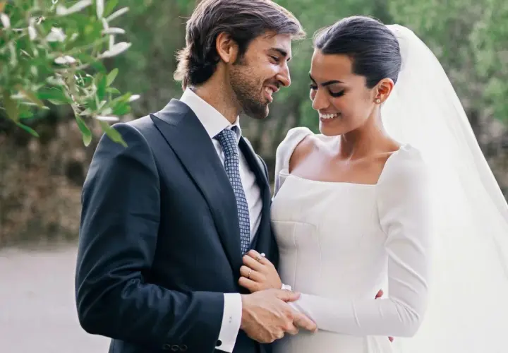 Marta Lozano y su esposo, radiantes en su boda, comparten una mirada cómplice y sonrisas. Él, con traje oscuro y corbata, la abraza tiernamente.