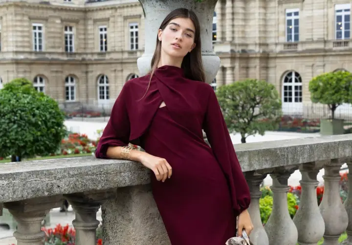 Mujer con un elegante vestido granate, apoyada en una barandilla de piedra, con un edificio histórico y jardines de fondo.