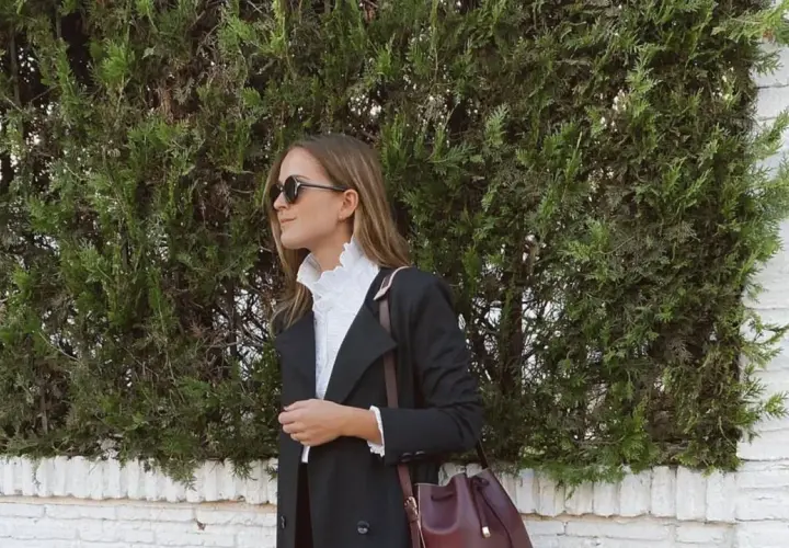 Mujer con elegante outfit para Semana Santa, abrigo negro, blusa blanca con volantes y bolso burdeos.