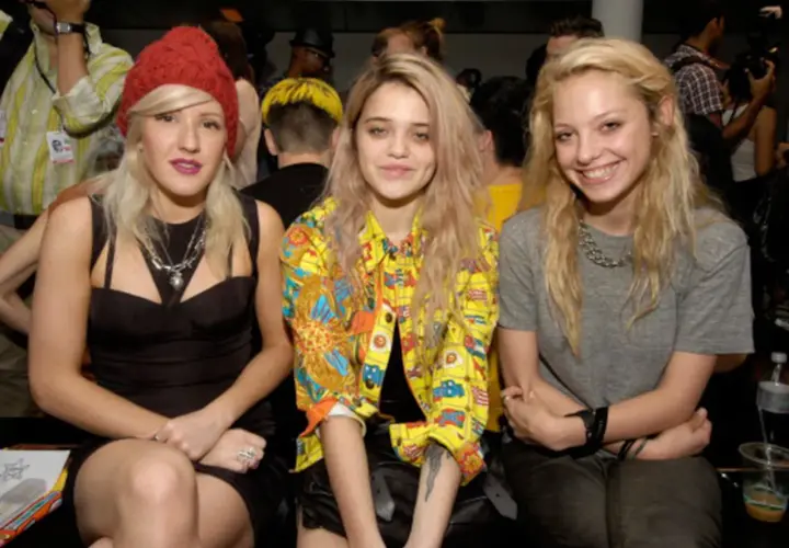 Tres jóvenes con un estilo que evoca el "heroin chic" posan juntas. Una lleva un gorro rojo, otra una chaqueta colorida y la tercera una camiseta gris.