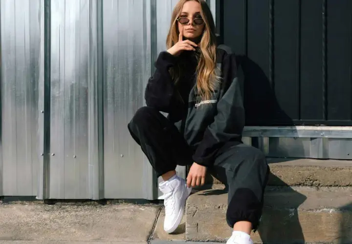 Chica con estilo moderno ropa, gafas de sol y zapatillas blancas, sentada en escalones de hormigón frente a una pared de metal.