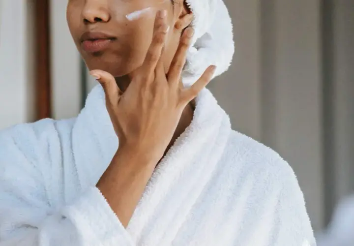 Mujer con bata blanca y toalla en la cabeza aplicándose crema en la mejilla, un gesto de cuidado para la piel con colágeno.