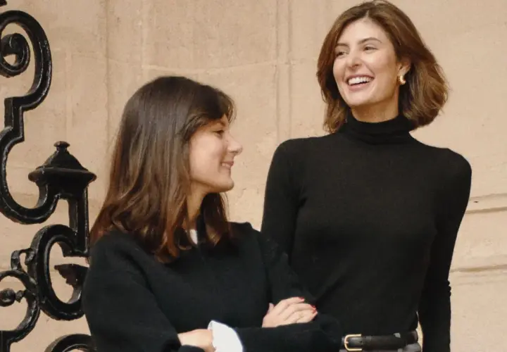María de la Orden y su amiga ríen juntas frente a una reja ornamental.