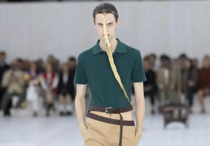 Enzo Vogrincic, modelo con un polo verde, pantalones caqui y un adorno dorado en la cara, desfila ante un público difuminado.