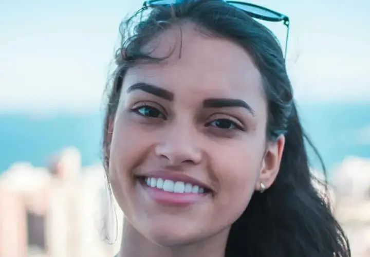 Sonrisa radiante de una joven con rostro ovalado, cabello oscuro y gafas de sol en la cabeza. Fondo de ciudad y mar.