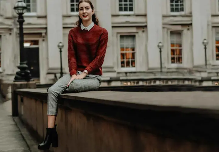 Chica con estilos de ropa modernos: jersey de punto rojo, camisa blanca, pantalones de cuadros y botines negros, sentada frente a un edificio clásico.