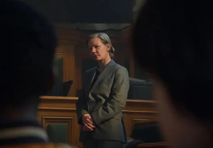 Sandra Hüller, con traje gris, se mantiene firme en una sala de audiencias, con las manos entrelazadas, mientras la cámara se enfoca en su rostro pensativo.