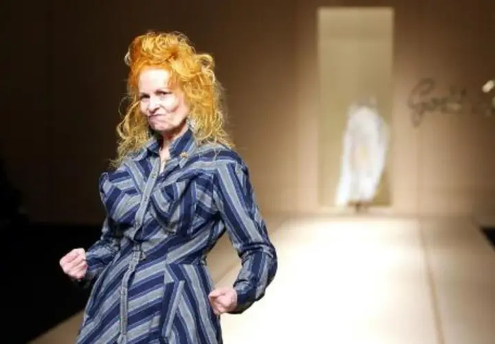 Vivienne Westwood, con su icónico cabello naranja, posa con energía en la pasarela de Westwood, luciendo un vestido a rayas azules y grises.