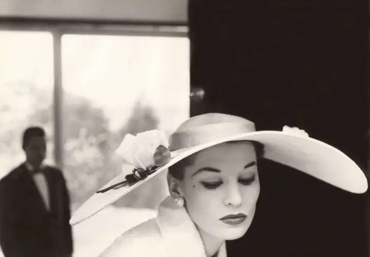 Mujer con sombrero de ala ancha y rosa, estilo Helmut Newton. Un hombre en traje con pajarita al fondo.