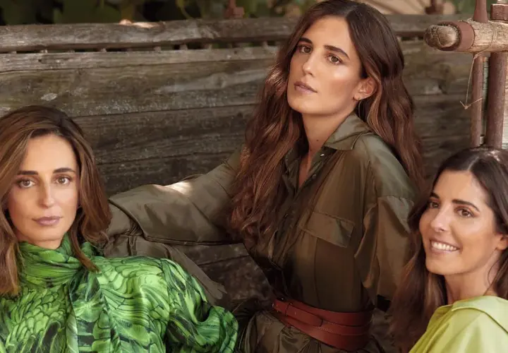 Belen Corsini y dos mujeres más, vestidas con ropa de moda, posan sentadas en un banco de madera rústico.