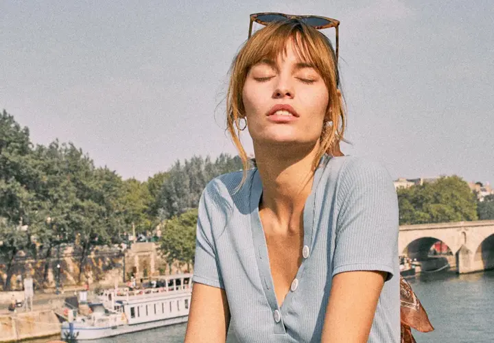 Mujer con gafas de sol en la cabeza y un pañuelo con estampado vichy, disfrutando del sol junto a un río con un barco y un puente al fondo.