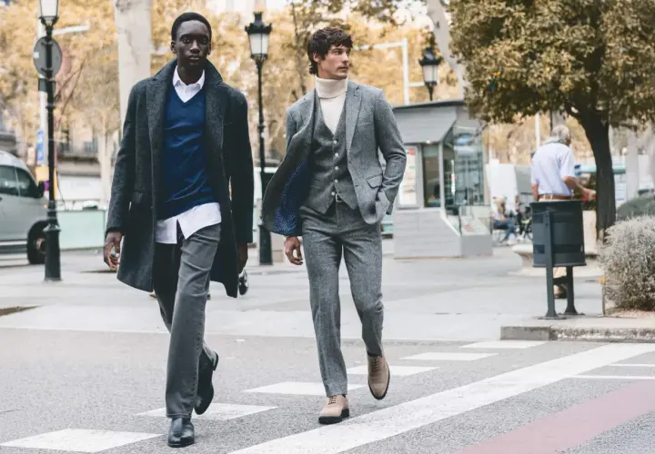 Dos hombres cruzan una calle, uno con abrigo y jersey azul, el otro con traje gris. Representan la moda slow, atemporal y consciente.