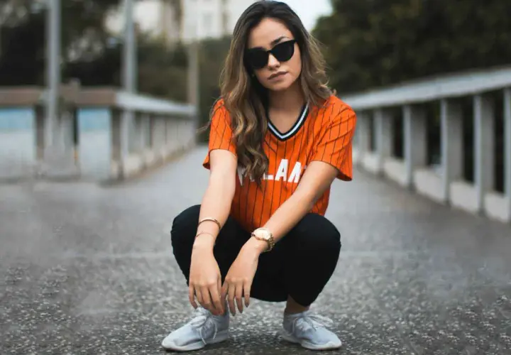 Chica con gafas de sol y camiseta naranja a rayas, agachada en un puente. Su atuendo es muy comfy y moderno.