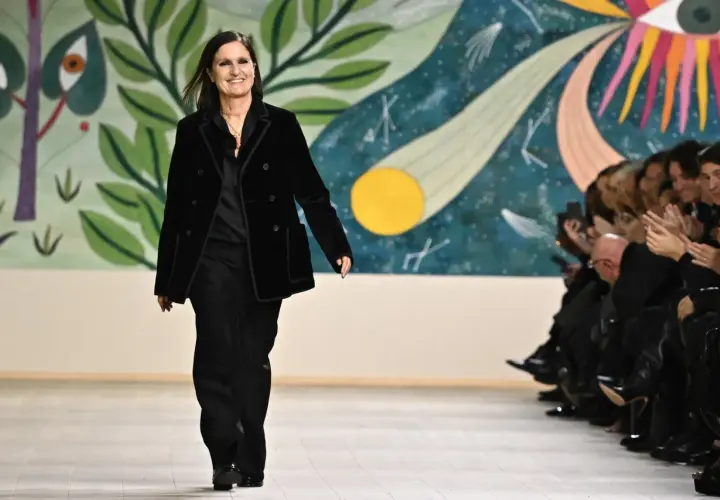 Maria Grazia Chiuri camina en la pasarela con un traje negro, sonriendo ante el público aplaudiendo.