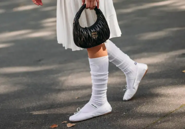 Piernas con bailarinas blancas y calcetines blancos arrugados, un bolso negro Miu Miu en la mano.