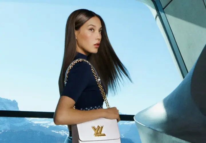 Eileen Gu, con su bolso Louis Vuitton, posa con elegancia frente a un paisaje montañoso y un cielo azul.
