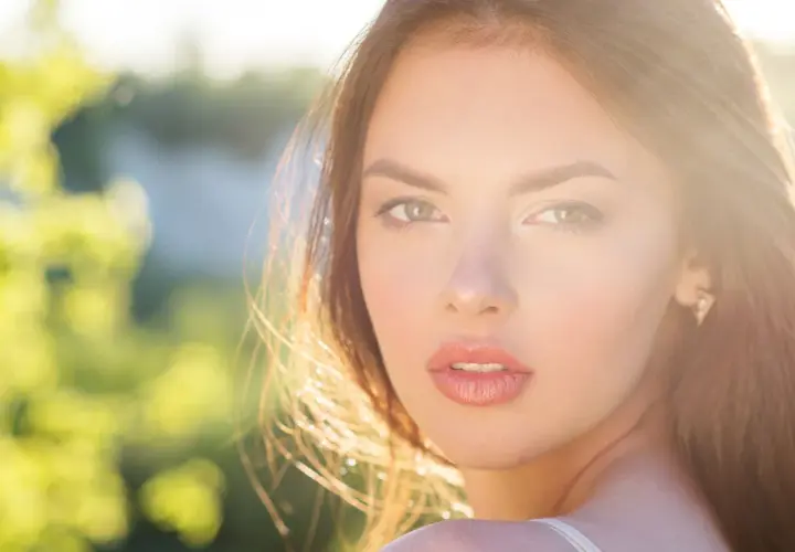 Una joven con un brillo natural, un verdadero glow up, mira a la cámara con el sol iluminando su rostro y el fondo verde desenfocado.
