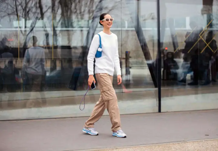 Mujer sonriente camina con un suéter blanco y un pantalón beige de carga, luciendo gafas de sol blancas y un bolso azul.