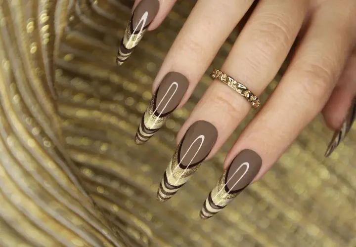 Uñas marrones tendencia con diseño de líneas doradas y brillo. Un anillo dorado adorna el dedo.