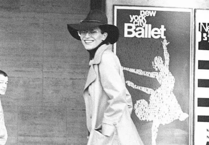 Elsa Peretti, con sombrero y gafas, sonríe frente a un cartel del New York City Ballet.