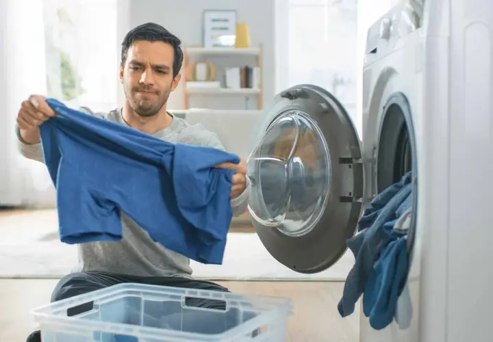 Hombre mira con preocupación una camiseta azul, parece que la viscosa encoge. La lavadora está abierta con ropa dentro.