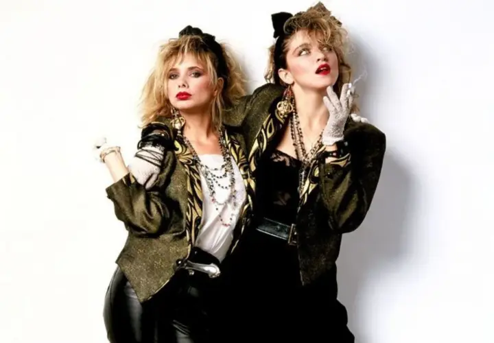 Dos mujeres posan con vestimenta años 80: chaquetas llamativas, guantes de encaje, collares largos y peinados voluminosos.