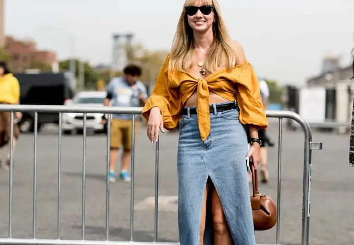 Mujer sonriente con un outfit falda vaquera larga, top amarillo anudado y bolso marrón.