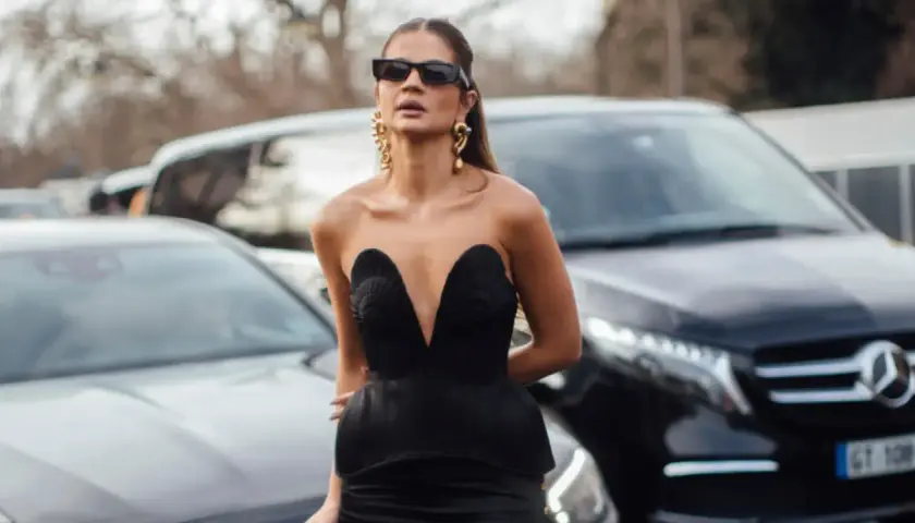Mujer con gafas de sol y pendientes dorados, vistiendo de negro un escotado vestido con detalles de conchas.