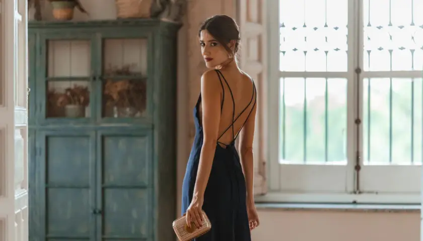 Mujer con un elegante vestido azul marino, perfecto para combinar en una boda. Lleva un bolso dorado y mira por encima del hombro.