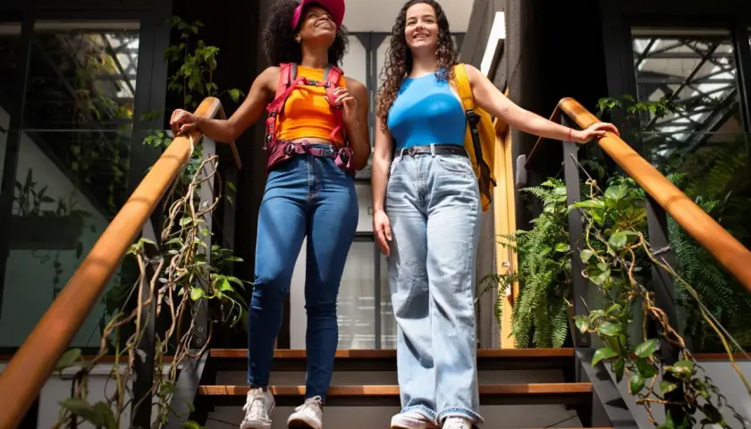 Dos amigas con estilo y2k, una con top naranja y gorra rosa, la otra con top azul, bajan unas escaleras rodeadas de plantas.