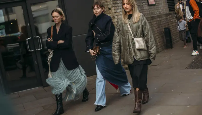 Tres mujeres caminan por la calle, cada una con un estilo único. La de la izquierda lleva unas **botas** negras altas y una falda de flecos. La del medio, un conjunto vaquero y unas botas negras. La de la derecha, unas botas marrones y una chaqueta aco...