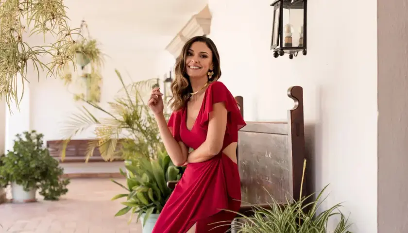 Mujer sonriente con un elegante outfit bautizo rojo, con detalles de corte y volantes, posando entre plantas y un banco de madera.