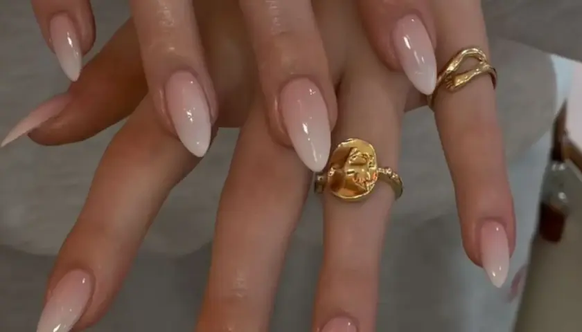 Manos con uñas baby boomer natural, un degradado suave de rosa a blanco. Dos anillos dorados adornan los dedos.