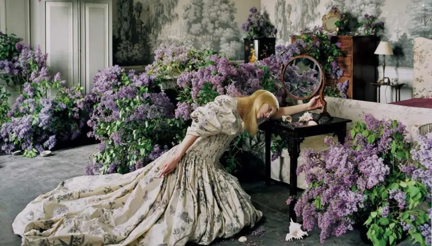 Una mujer rubia con un vestido estampado cae sobre un montón de lilas, evocando la estética onírica de Tim Walker.