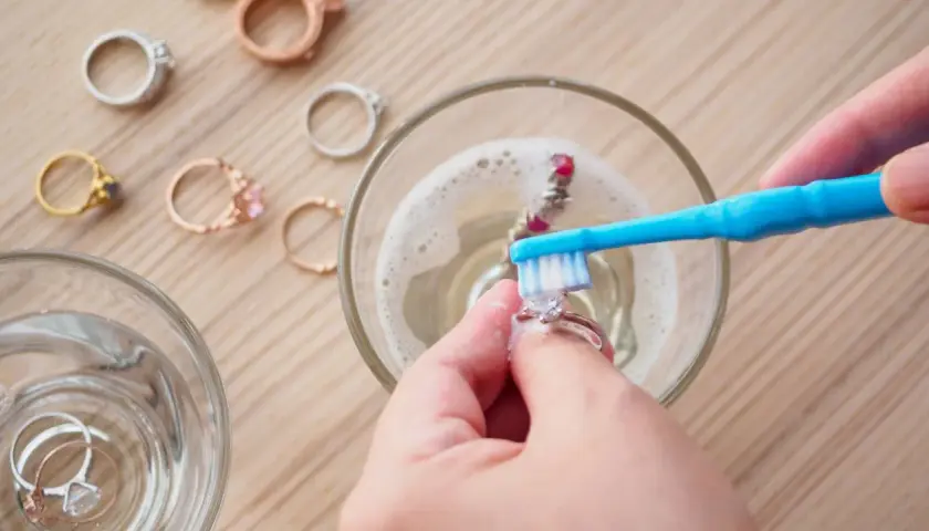 Manos limpian joyas con un cepillo azul en agua con espuma. Otros anillos esperan su turno.