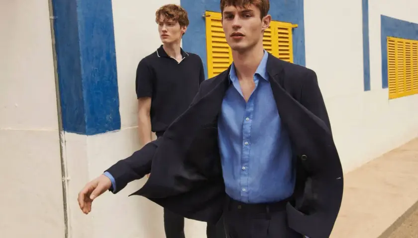 Dos hombres modelan un outfit oficina verano hombre: uno con chaqueta azul marino y camisa de lino, otro con polo oscuro.