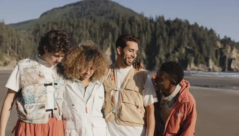 Grupo de amigos con estilo gorpcore en la playa, con chalecos utilitarios y chaquetas, disfrutando del paisaje costero.